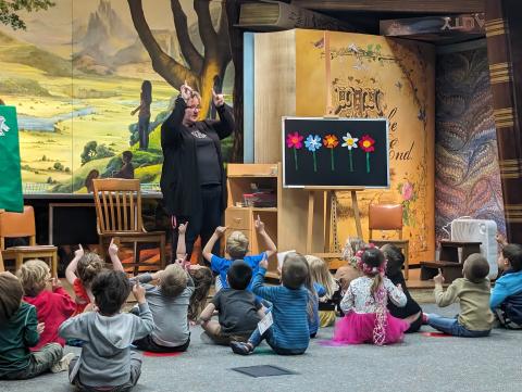 Miss Sharon at flannel board with children sitting on floor with arms up in the air.