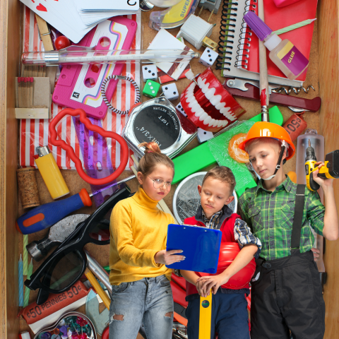 Three child engineers look at plans. A junk drawer in the background is filled with colorful ephemera.