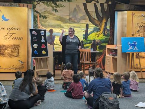 Miss Sharon at flannel board with children and adults sitting on floor with arms up in the air.