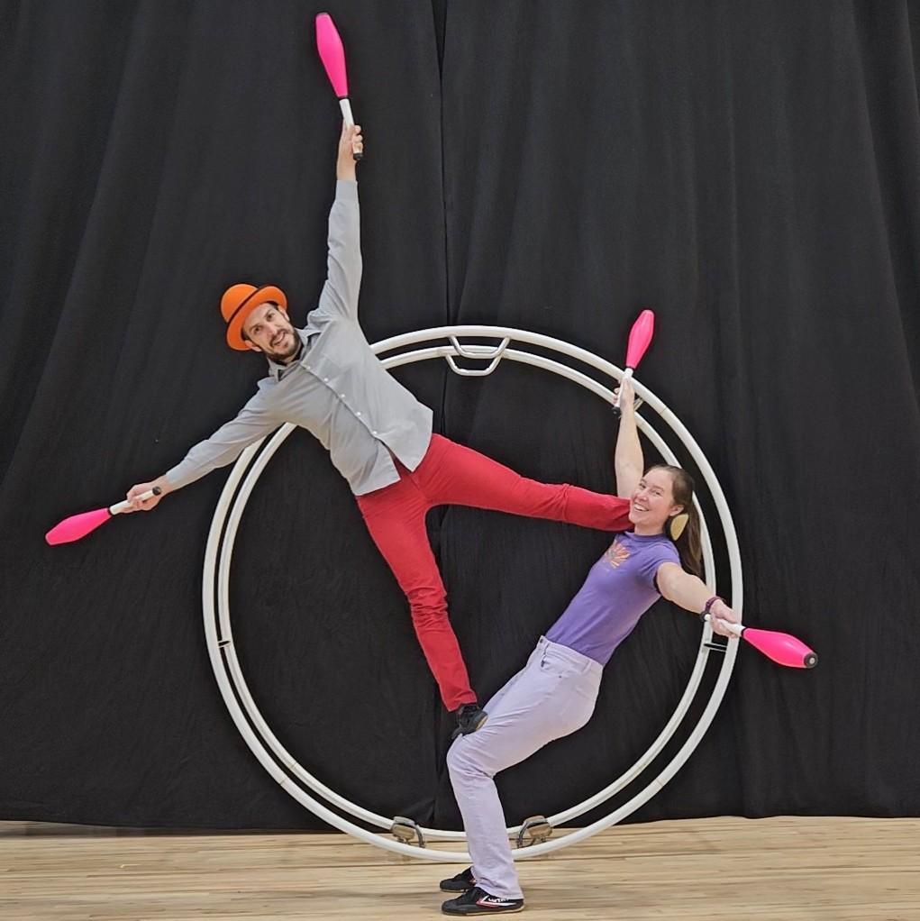 Circus performers hold juggling pins while balancing together on a German wheel.